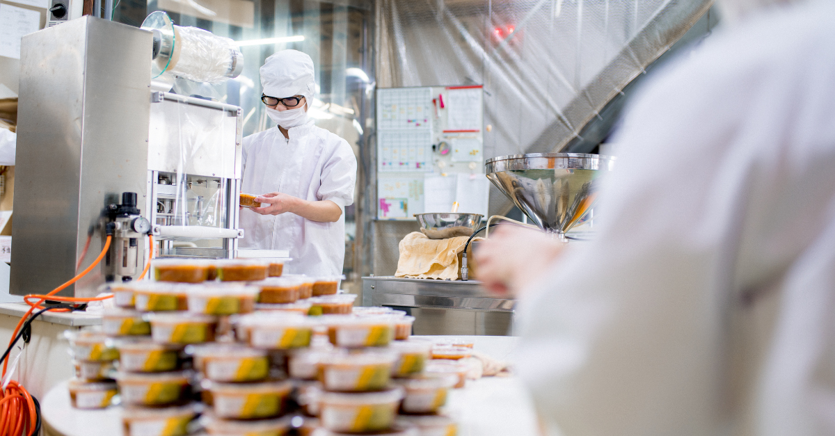 Person in lab coat and mask applying lids to food packages behind mound of packages